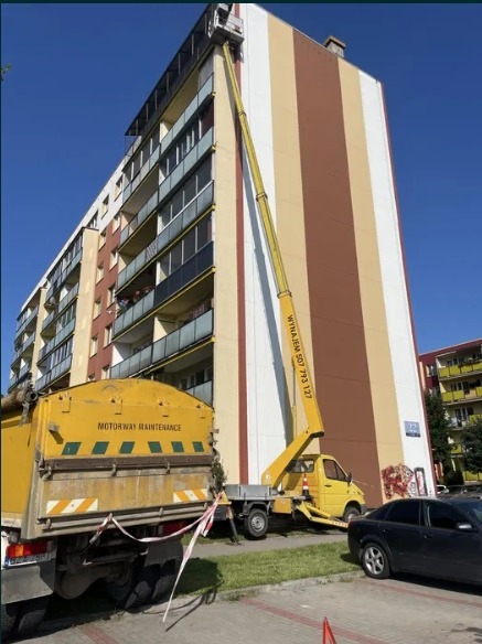 Żółty pojazd z wysięgnikiem na tle wysokiego bloku z pasami w odcieniach beżu i brązu. Na wysięgniku platforma robocza. Widoczny fragment pojazdu 'Motorway Maintenance'.