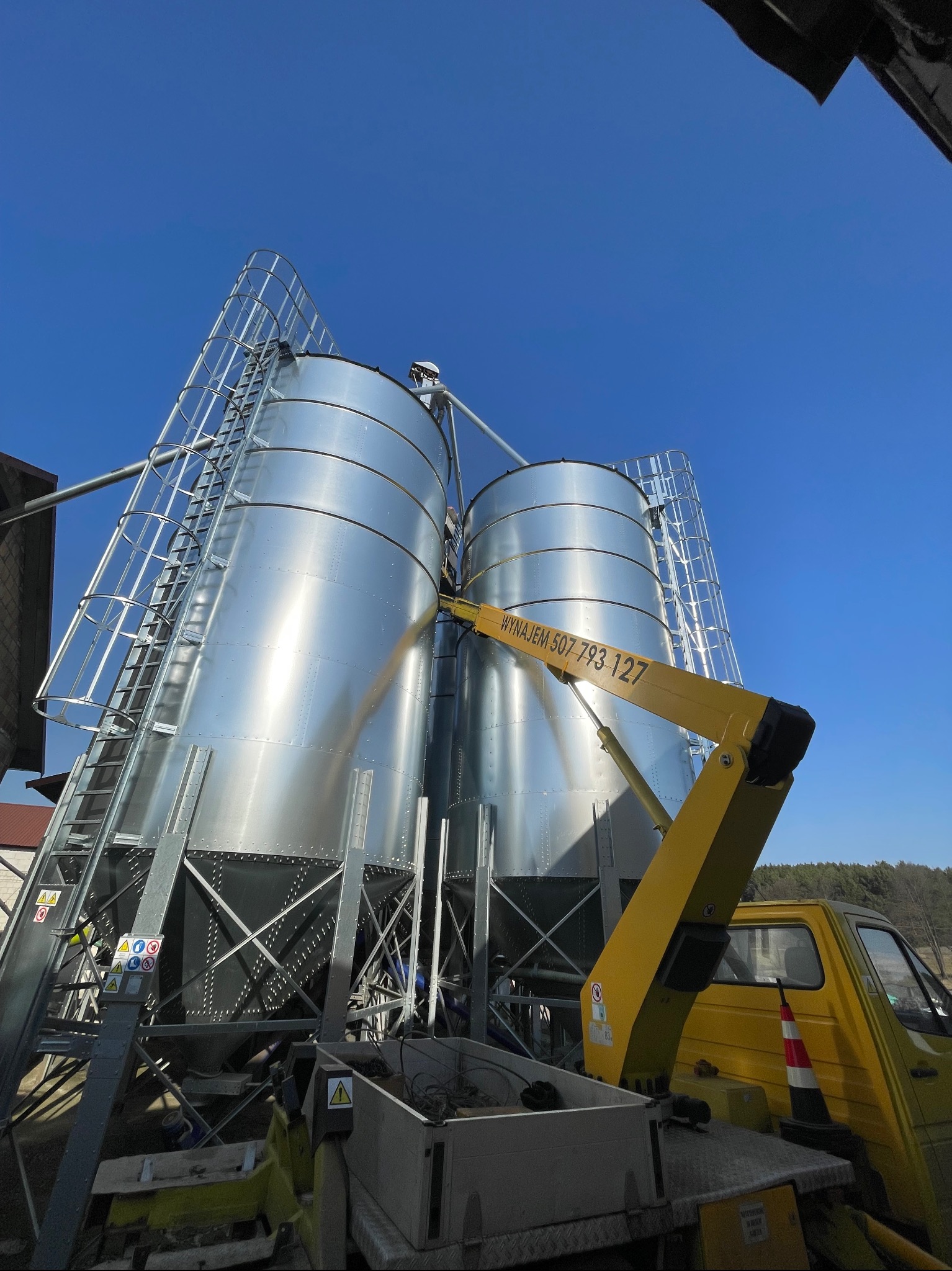 Dwa srebrne silosy zbożowe z drabinkami, oświetlone słońcem, z żółtym ramieniem podnośnika koszowego z napisem 'Wynajem 507 793 127' na tle błękitnego nieba.