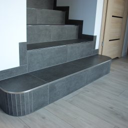 Jakub Frączek Wykończenia Wnętrz - Schody wykończone szarą płytką z metalowymi listwami, dopasowane do podłogi z paneli. Widoczne drzwi wewnętrzne z jasnego drewna.