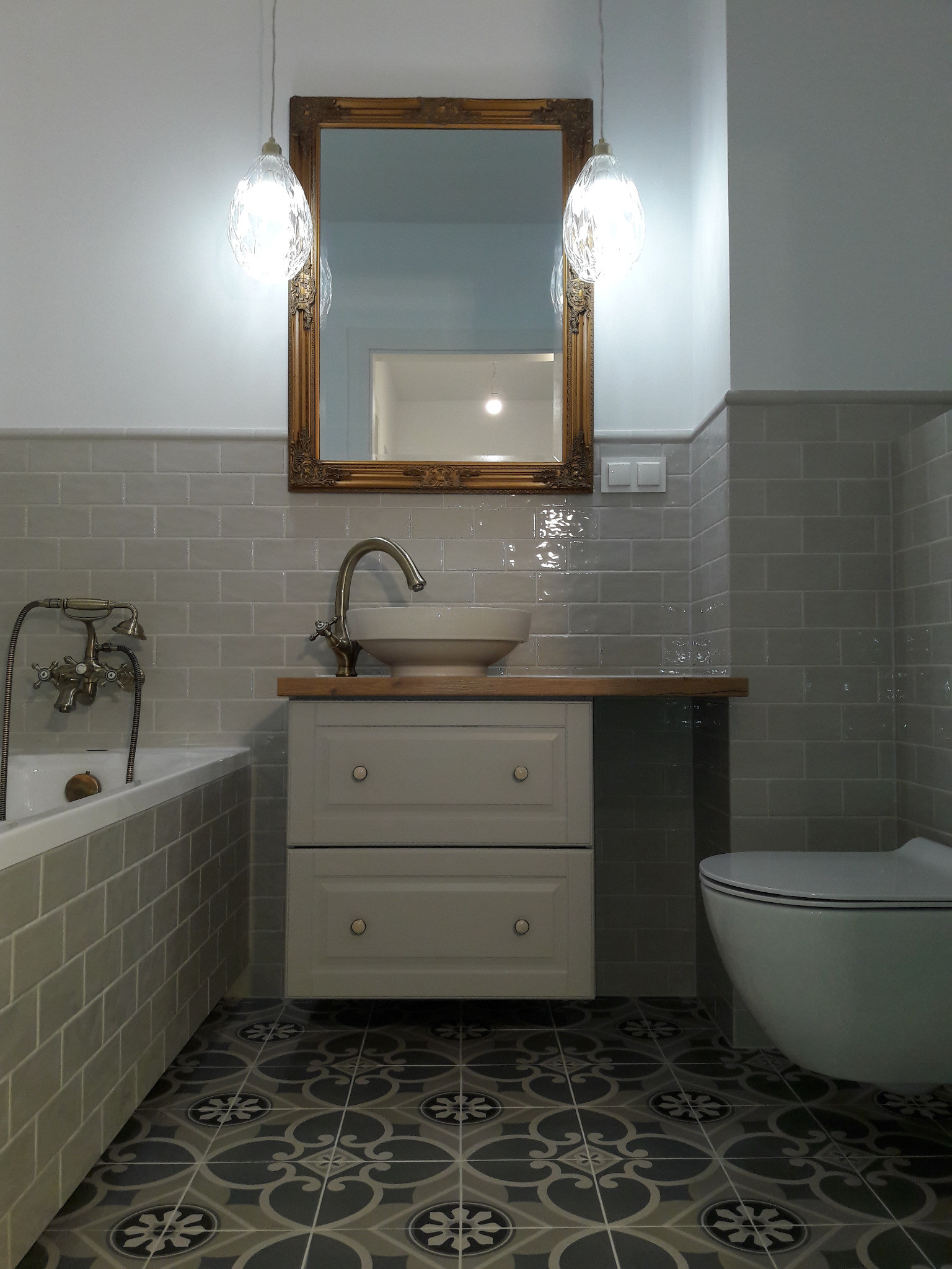 Stylowa łazienka z szarymi płytkami, wzorzystą podłogą, umywalką na szafce z drewnianym blatem i złotym lustrem. Dwie wiszące lampy dodają elegancji.