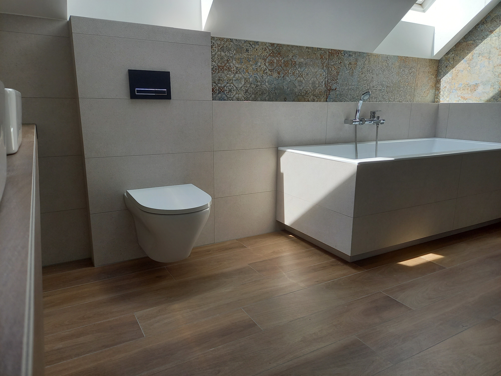 Nowoczesna łazienka na poddaszu z drewnopodobną podłogą, beżowymi płytkami i wzorzystym pasem dekoracyjnym. Wanna obudowana płytkami, podwieszana toaleta.