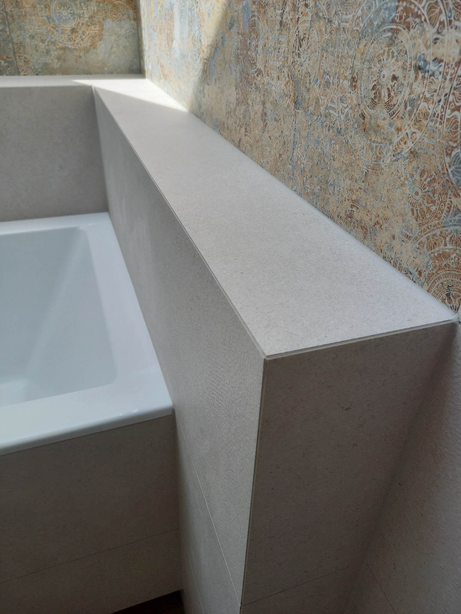 Obudowa wanny z szarej, teksturowanej płyty, z dekoracyjną ścianą w tle. Widoczny fragment białej wanny. Kątowe ujęcie podkreśla precyzję wykonania.