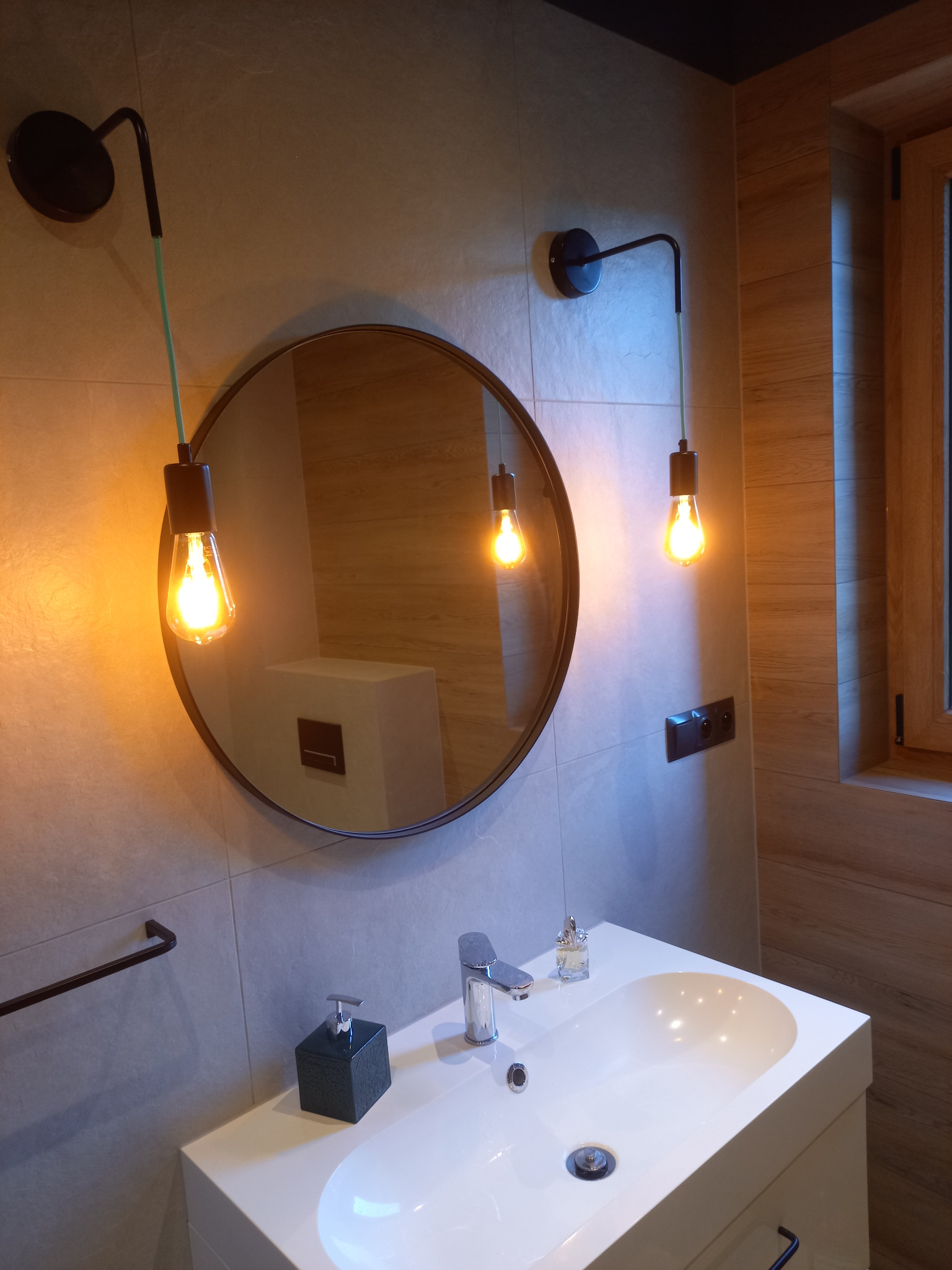 Nowoczesna łazienka z okrągłym lustrem, wiszącymi lampami z żarówkami Edisona i białą umywalką. Ściany wyłożone płytkami imitującymi kamień, drewniane wykończenie okna.