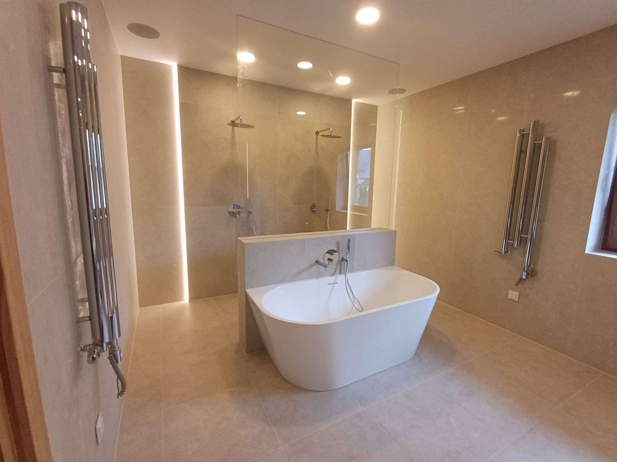 Nowoczesna łazienka z wolnostojącą wanną, przeszklonym prysznicem i grzejnikami drabinkowymi. Beżowe płytki, subtelne oświetlenie LED. Minimalistyczny design.