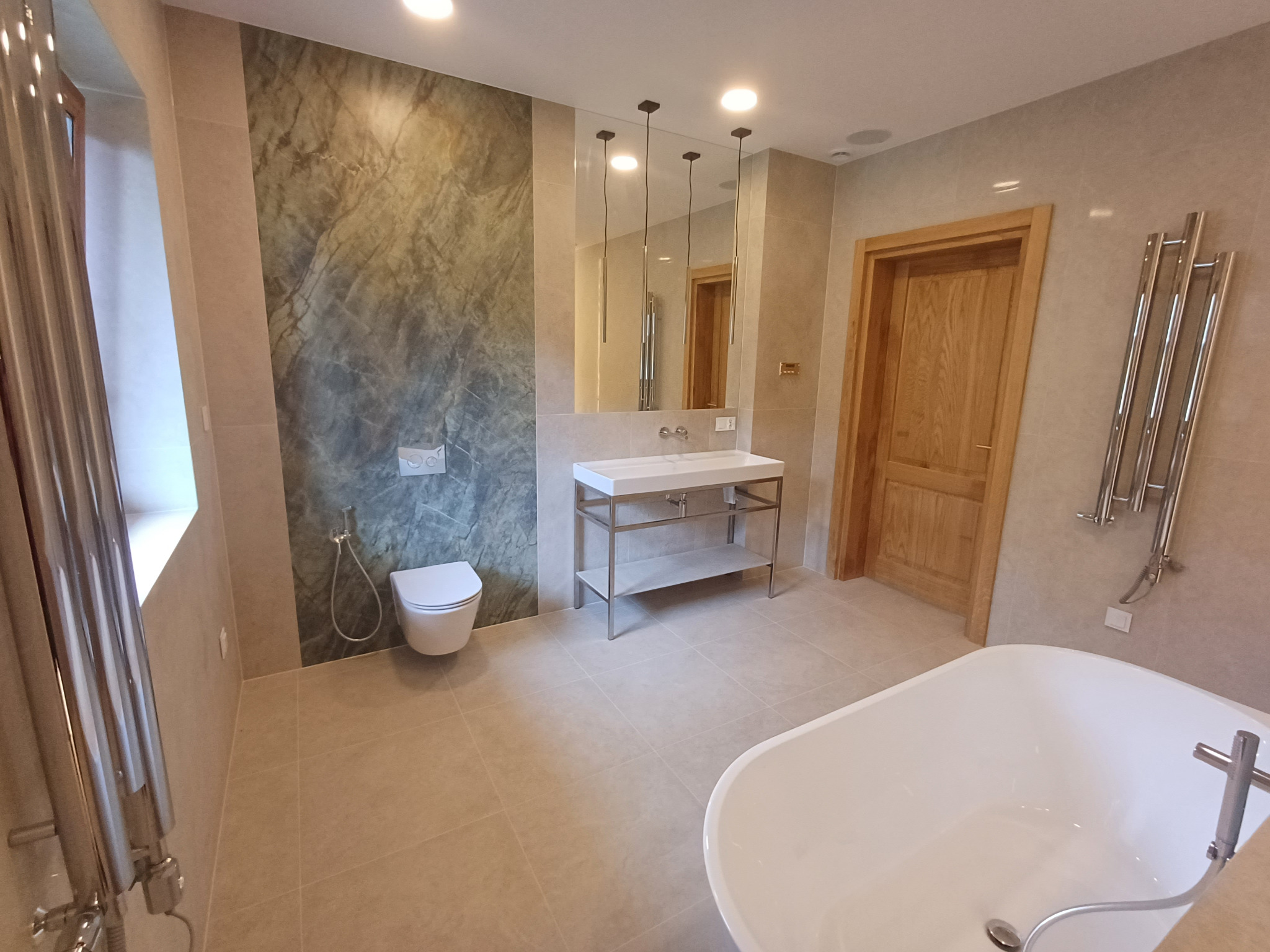 Nowoczesna łazienka z marmurową ścianą, wiszącą toaletą i umywalką na metalowym stelażu. Minimalistyczne oświetlenie i drewniane drzwi dodają elegancji.