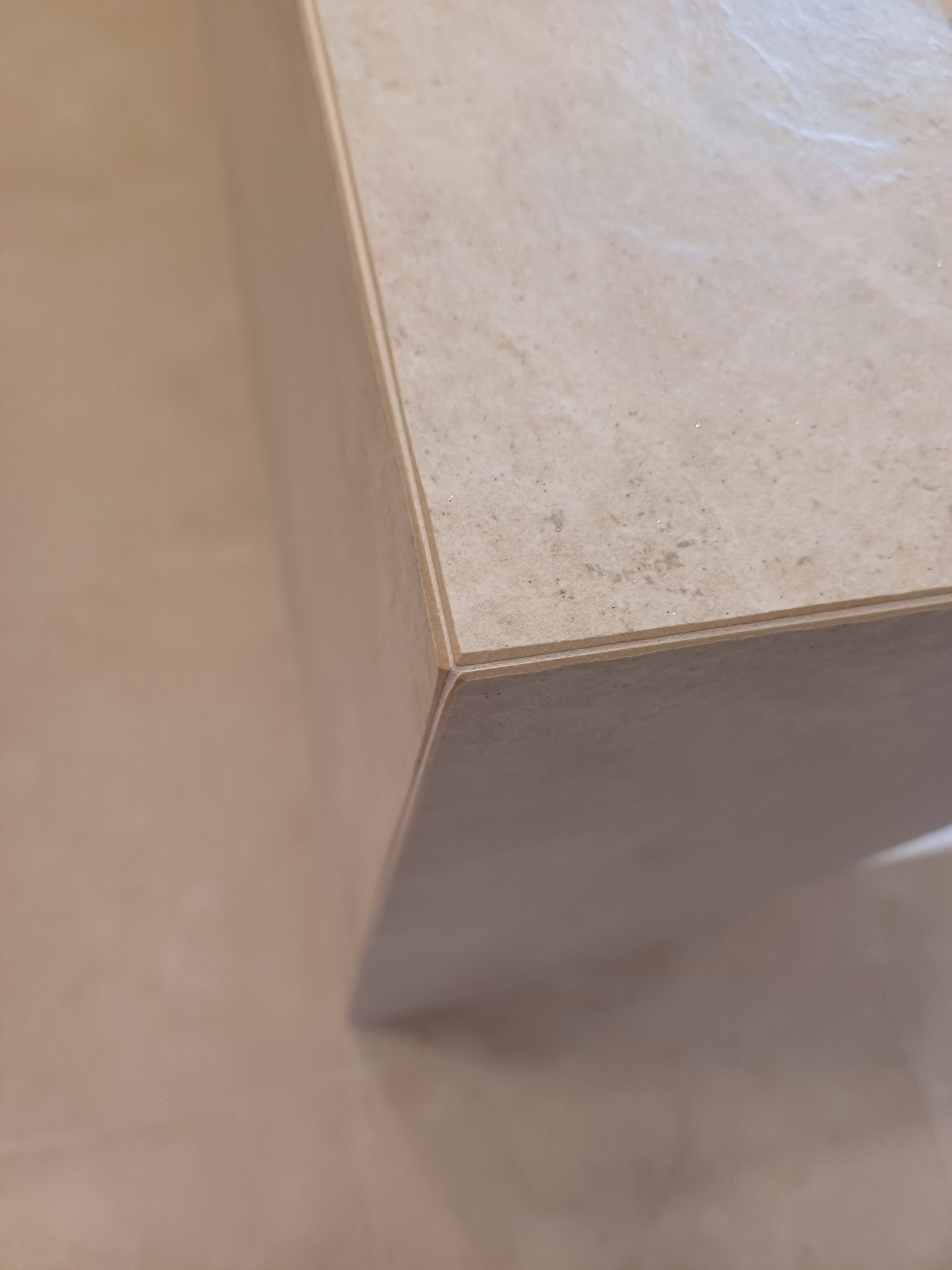 Beżowa, marmurkowa płyta ceramiczna, widoczny narożnik i krawędź. Delikatna faktura kamienia, ciepłe odcienie beżu. Minimalistyczny detal wykończenia wnętrza.