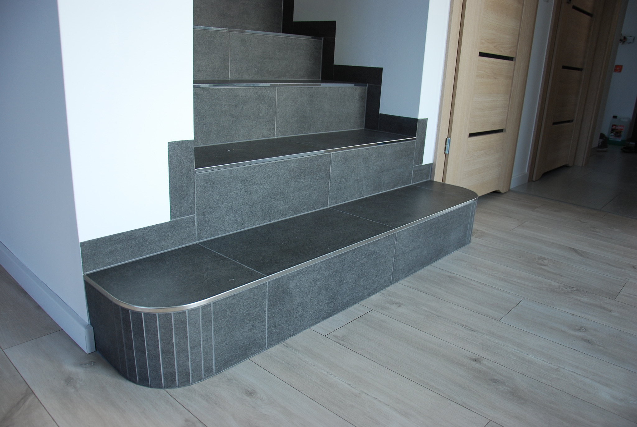 Schody wykończone szarą płytką z metalowymi listwami, dopasowane do podłogi z paneli. Widoczne drzwi wewnętrzne z jasnego drewna.