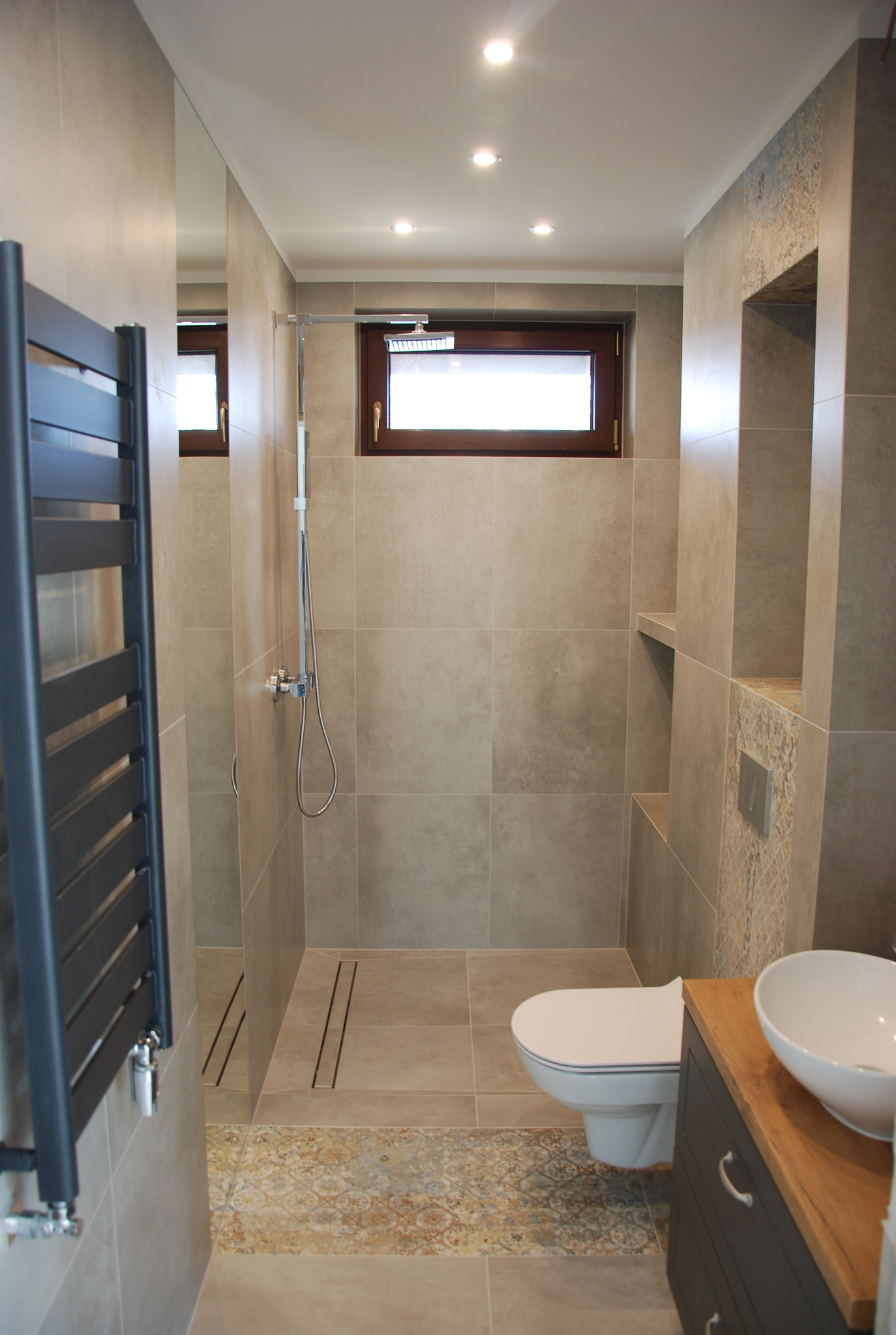 Nowoczesna łazienka z beżowymi płytkami, kabiną prysznicową typu walk-in, grzejnikiem drabinkowym i umywalką nablatową na drewnianym blacie. Widoczne okno i oświetlenie punktowe.