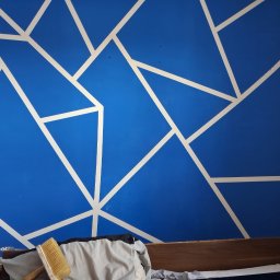 Niebieska ściana z geometrycznym wzorem w bieli, świeżo po malowaniu w Bystrzycy. Widoczny pędzel i fragment mebli, co sugeruje domowe otoczenie.