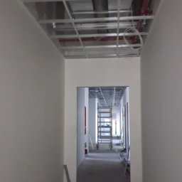 Korytarz w trakcie prac wykończeniowych: pomalowane ściany, widoczna konstrukcja sufitu podwieszanego i drabina w oddali. Minimalistyczne wnętrze w jasnych barwach.