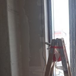 Ściana po szpachlowaniu, widoczna drabina malarska z narzędziami. Przygotowanie do dalszych prac wykończeniowych w pomieszczeniu z oknem.