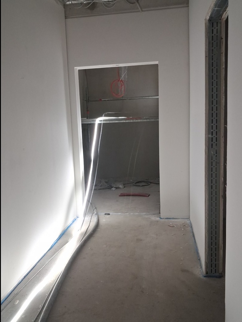 Wnętrze w trakcie prac wykończeniowych: gładkie, białe ściany, surowy beton na podłodze, widoczne kable i instalacje. Otwór drzwiowy z metalową listwą wykończeniową.