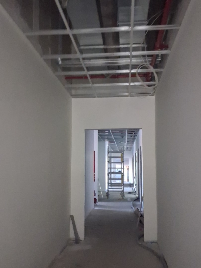Korytarz w trakcie prac wykończeniowych: pomalowane ściany, widoczna konstrukcja sufitu podwieszanego i drabina w oddali. Minimalistyczne wnętrze w jasnych barwach.