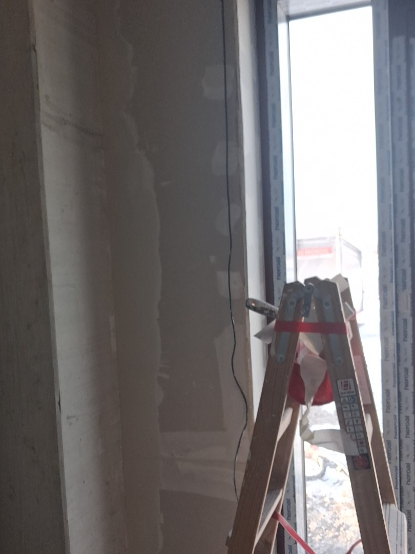 Ściana po szpachlowaniu, widoczna drabina malarska z narzędziami. Przygotowanie do dalszych prac wykończeniowych w pomieszczeniu z oknem.