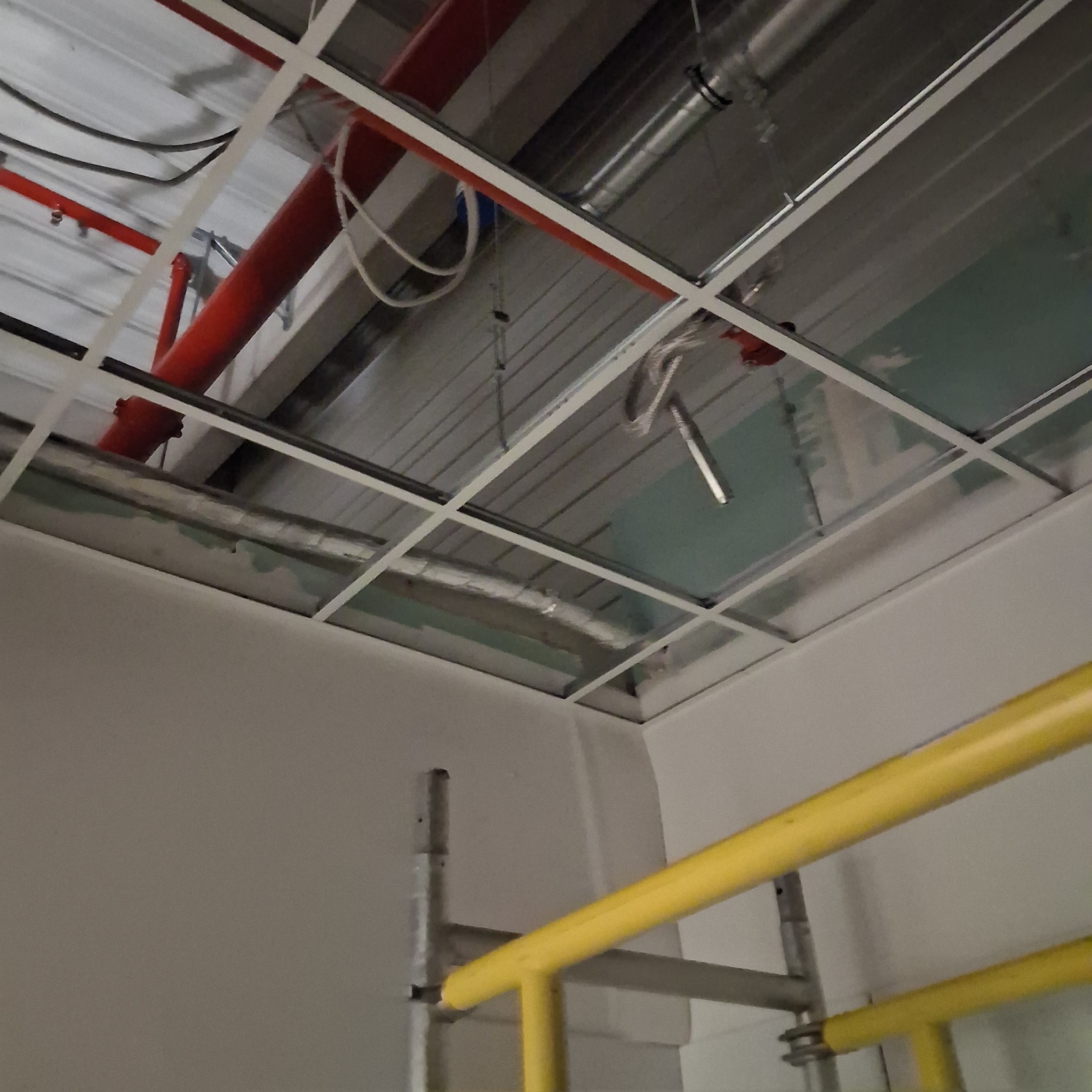 Montaż sufitu podwieszanego z widocznymi instalacjami i fragmentem rusztowania. Biała kratownica, czerwone rury i srebrne przewody nad płytami.