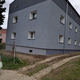 PRO-BUD JÓZEF KUNASZYK - Dwukolorowa elewacja budynku po remoncie, z widocznymi oknami i rynną spustową. Świeżo pomalowane ściany w odcieniach szarości. Widok z poziomu chodnika.