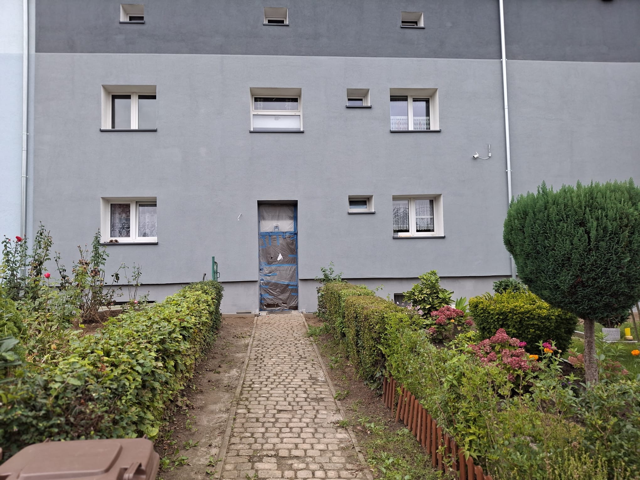 Szary budynek po ociepleniu z zabezpieczonym wejściem, widok z ogrodu z kamienną ścieżką. Świeżo otynkowana elewacja z oknami o różnych rozmiarach.