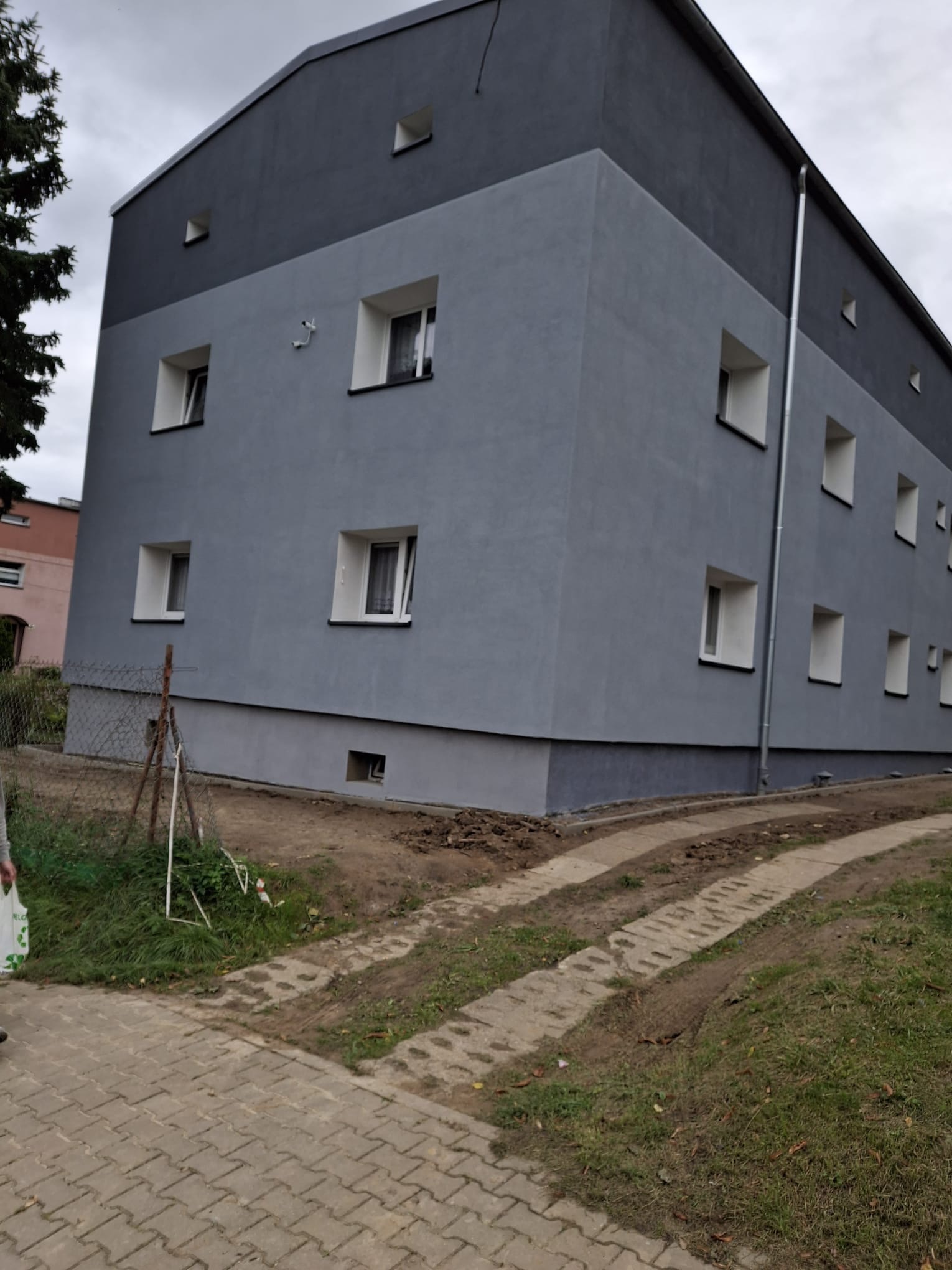 Dwukolorowa elewacja budynku po remoncie, z widocznymi oknami i rynną spustową. Świeżo pomalowane ściany w odcieniach szarości. Widok z poziomu chodnika.