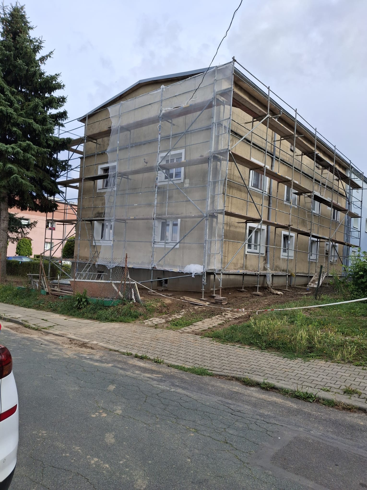Budynek wielorodzinny w trakcie prac elewacyjnych, osłonięty siatką rusztowaniową. Widoczne rusztowania, okna i fragment drzewa. Ujęcie z poziomu ulicy.