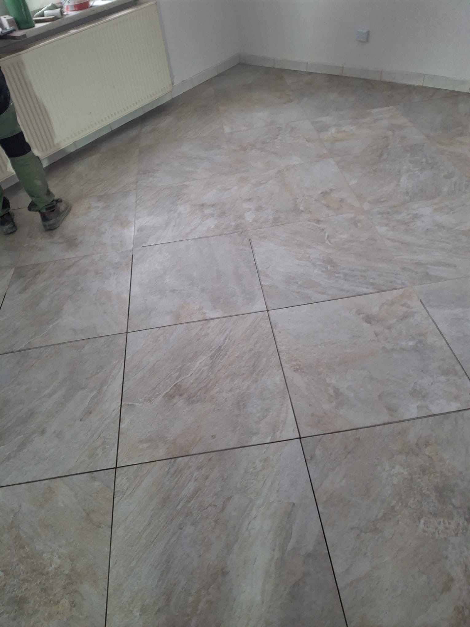 Ułożona podłoga z płytek imitujących kamień w jasnych odcieniach beżu i szarości. Widoczny fragment grzejnika i osoba w roboczym ubraniu.