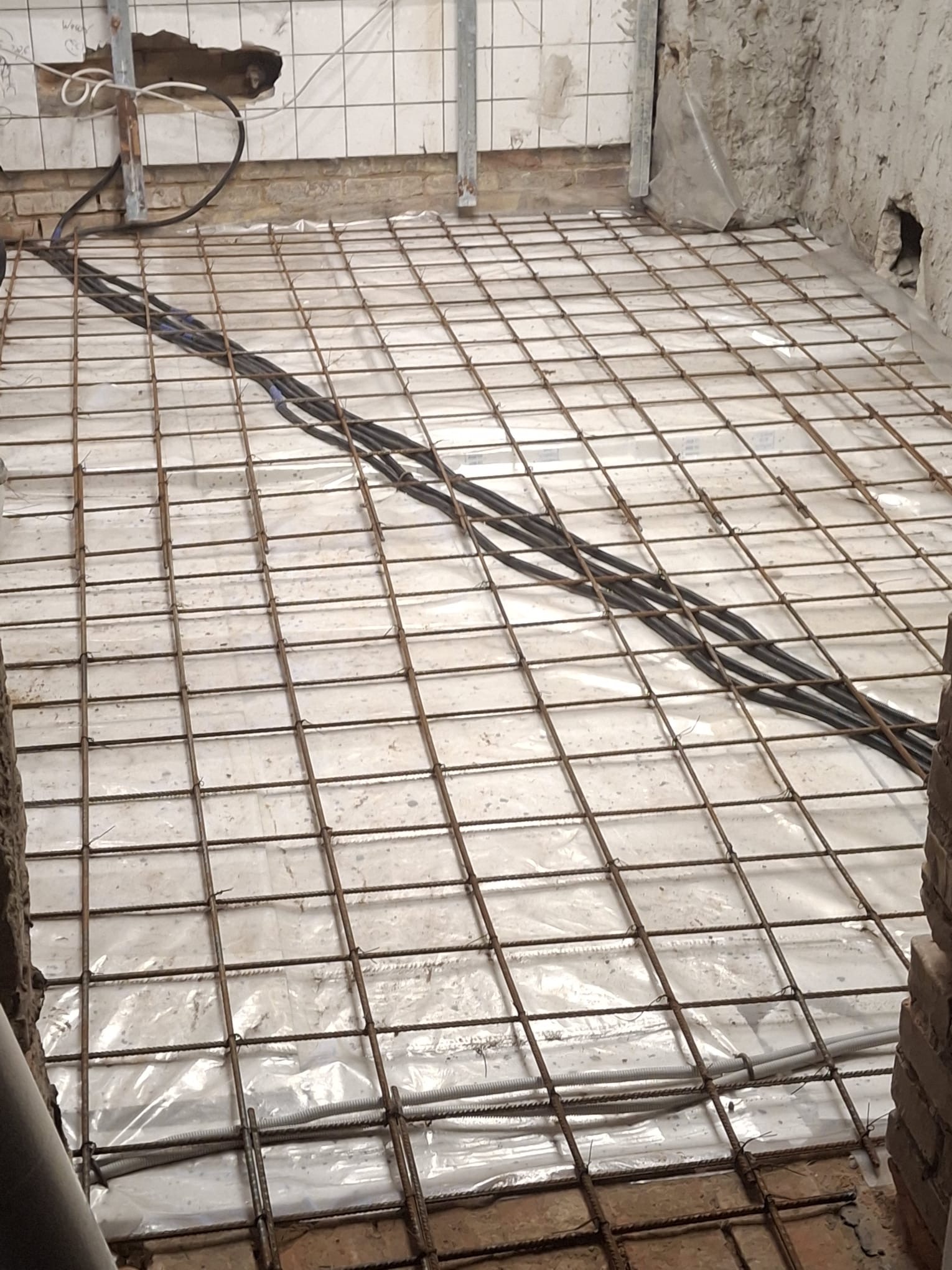 Przygotowanie podłoża: siatka zbrojeniowa i kable na folii w surowym wnętrzu. Widoczne fragmenty ścian z cegły i płytek, otwór w ścianie. Etap prac przed wylewką.
