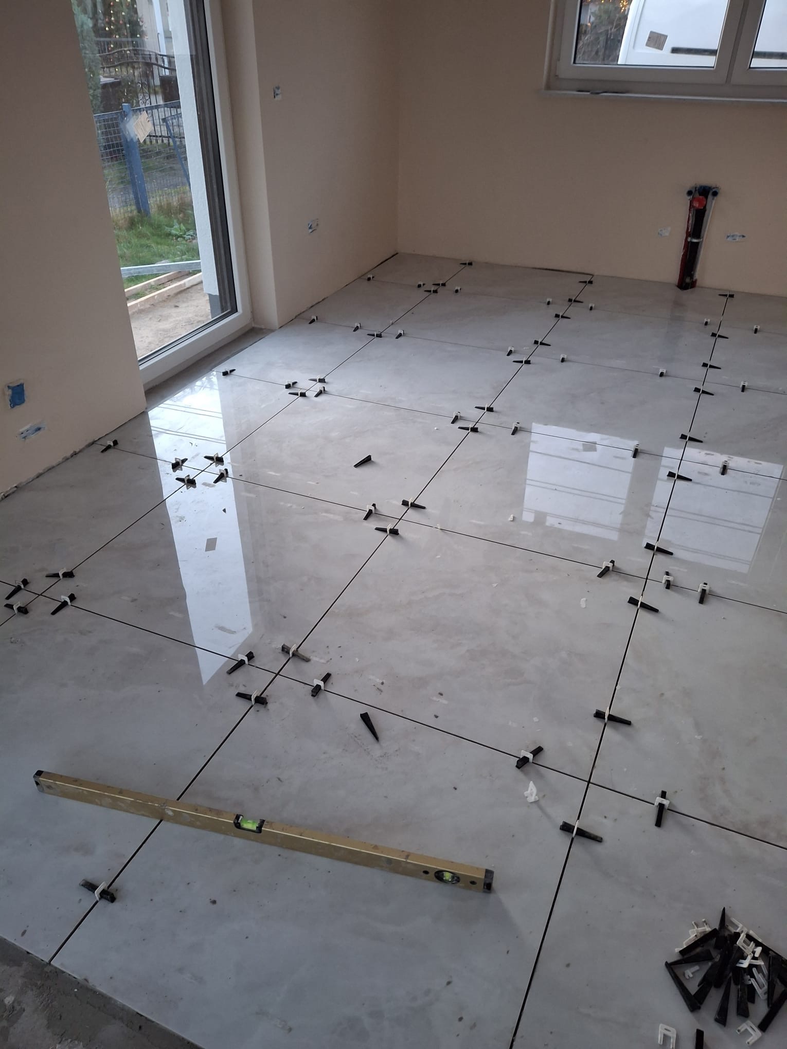 Układanie błyszczących, szarych płytek podłogowych z użyciem poziomicy i klipsów dystansowych. Widoczne okno i fragment ściany. Praca w toku.