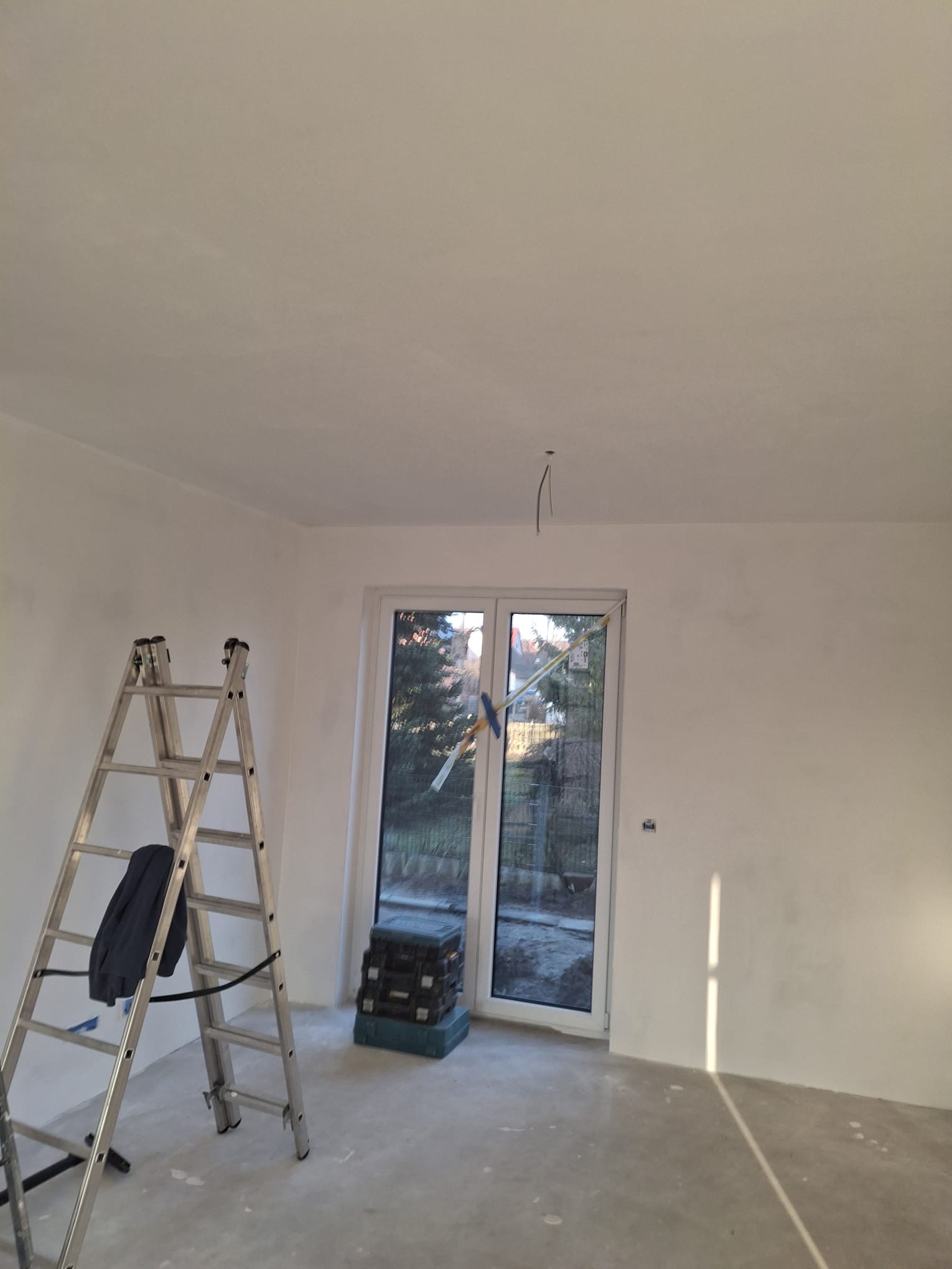 Białe, świeżo wyszpachlowane ściany w pokoju z oknem, drabina aluminiowa z ubraniem i skrzynki narzędziowe. Widoczne ślady szpachlowania na ścianach i suficie.