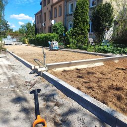 HYDRO ABS mgr inż. Bartosz Siwiec - Przygotowanie podłoża pod kostkę brukową w Świdwinie. Obrzeża ułożone, teren wyrównany, zagęszczarka widoczna w tle. Łopata wbita w ziemię na pierwszym planie.