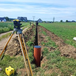 HYDROABS - Wykop pod instalacje w terenie wiejskim, widoczny pomarańczowy rurociąg i niwelator Leica na statywie. W tle domy i błękitne niebo.