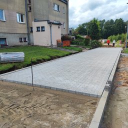 HYDROABS - Ułożona, szara kostka brukowa tworząca chodnik przed budynkiem mieszkalnym. Częściowo utwardzony teren z widocznymi śladami opon. Paleta kostki brukowej w tle.
