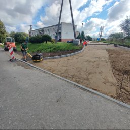 HYDROABS - Prace ziemne: zagęszczarka w akcji na przygotowanym podłożu, obok ułożone krawężniki. W tle budynek mieszkalny i maszyna budowlana. Etap przygotowania podłoża pod kostkę.