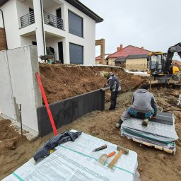 HYDROABS - Montaż paneli ogrodzeniowych na nierównym terenie, z koparką w tle. Widoczne narzędzia i materiały budowlane. Praca w toku, dom w tle.