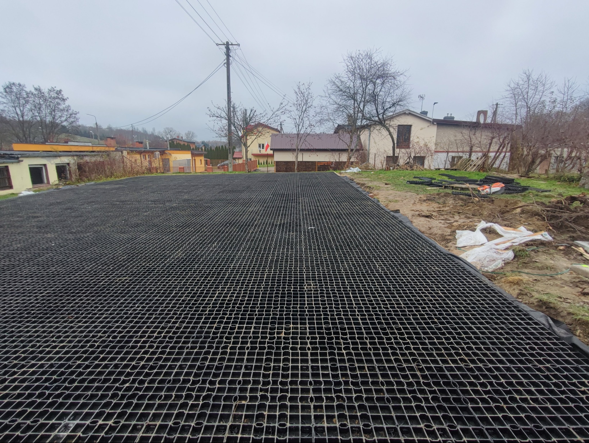 Ułożona, czarna geokrata na przygotowanym terenie. Widoczne budynki w tle i elementy krajobrazu. Etap przygotowania podłoża pod nawierzchnię.