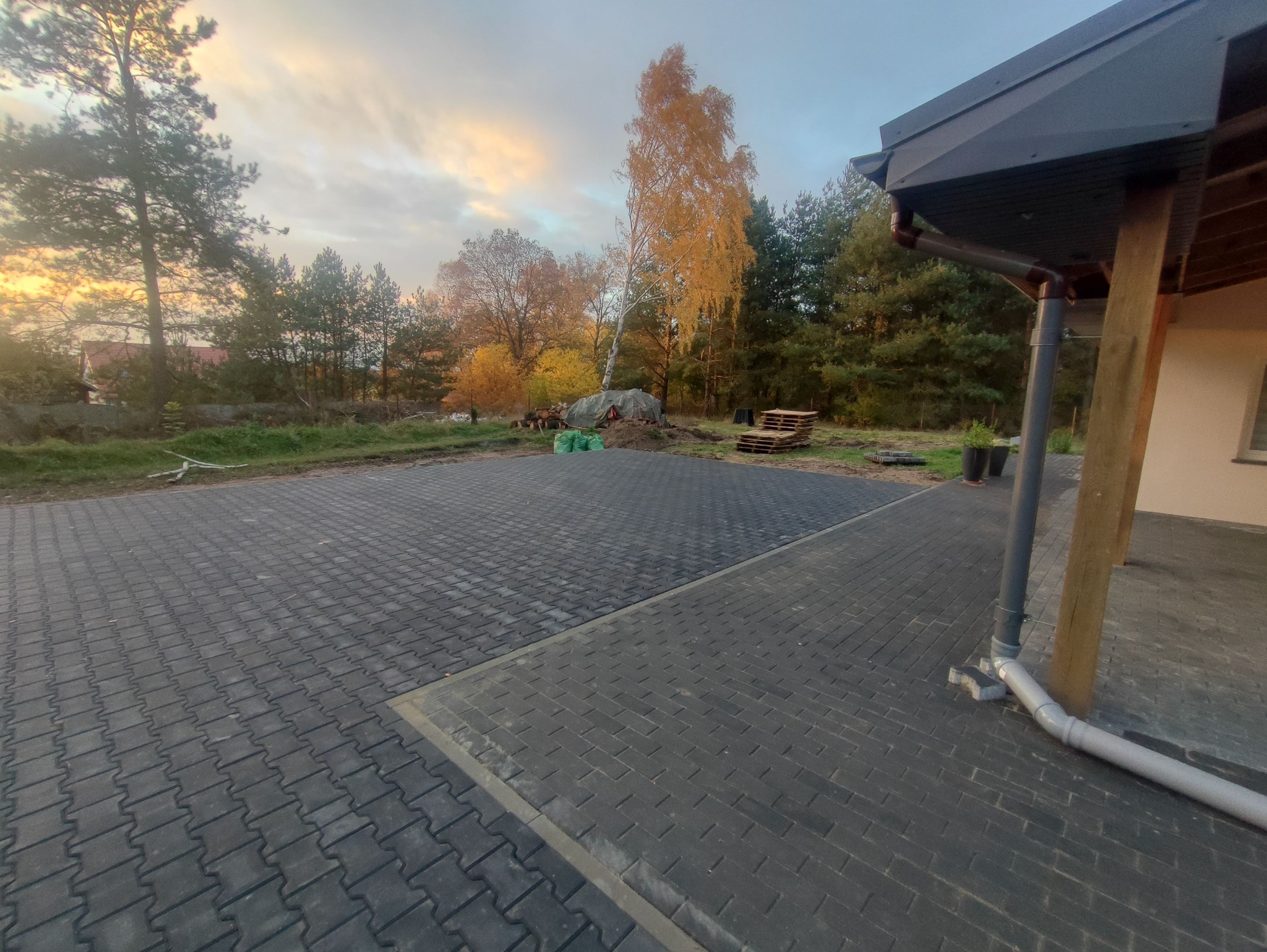 Ukończona nawierzchnia z ciemnej kostki brukowej na posesji, widok z poziomu gruntu, z fragmentem domu i jesiennym krajobrazem w tle. Widoczne palety i worki.