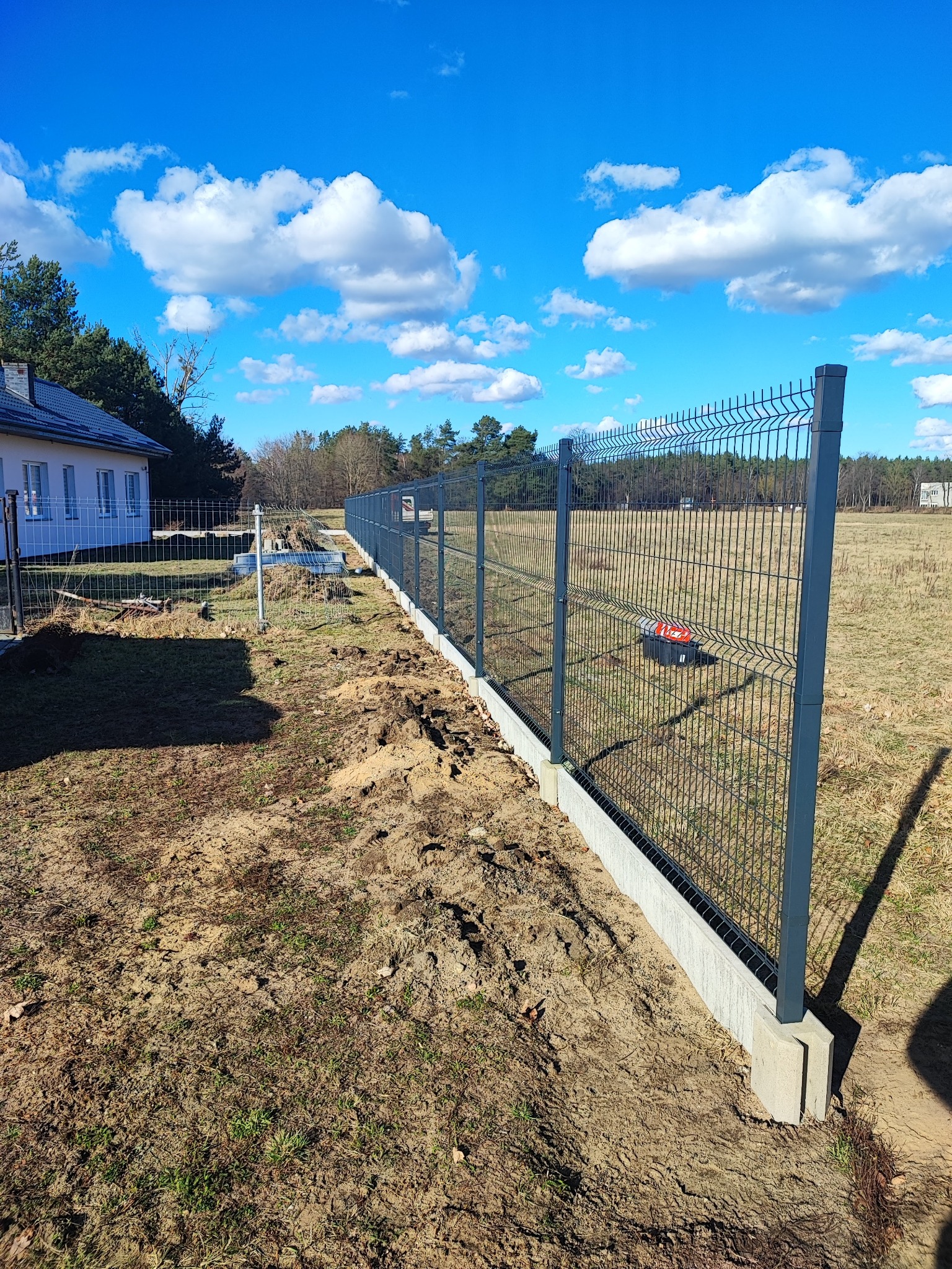 Nowoczesne ogrodzenie panelowe z podmurówką betonową, widok z boku na tle błękitnego nieba z chmurami. Częściowo zagospodarowany teren wokół ogrodzenia.