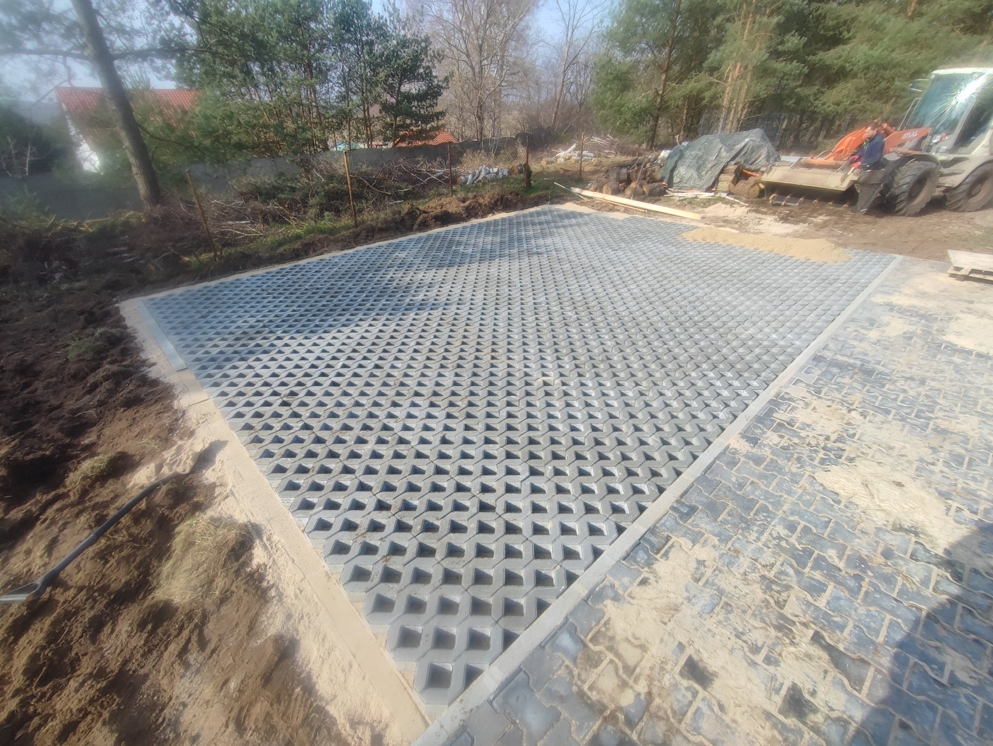 Ułożona szara ażurowa kostka brukowa na przygotowanym podłożu, obok tradycyjna kostka. W tle drzewa i maszyna budowlana. Widok z góry.