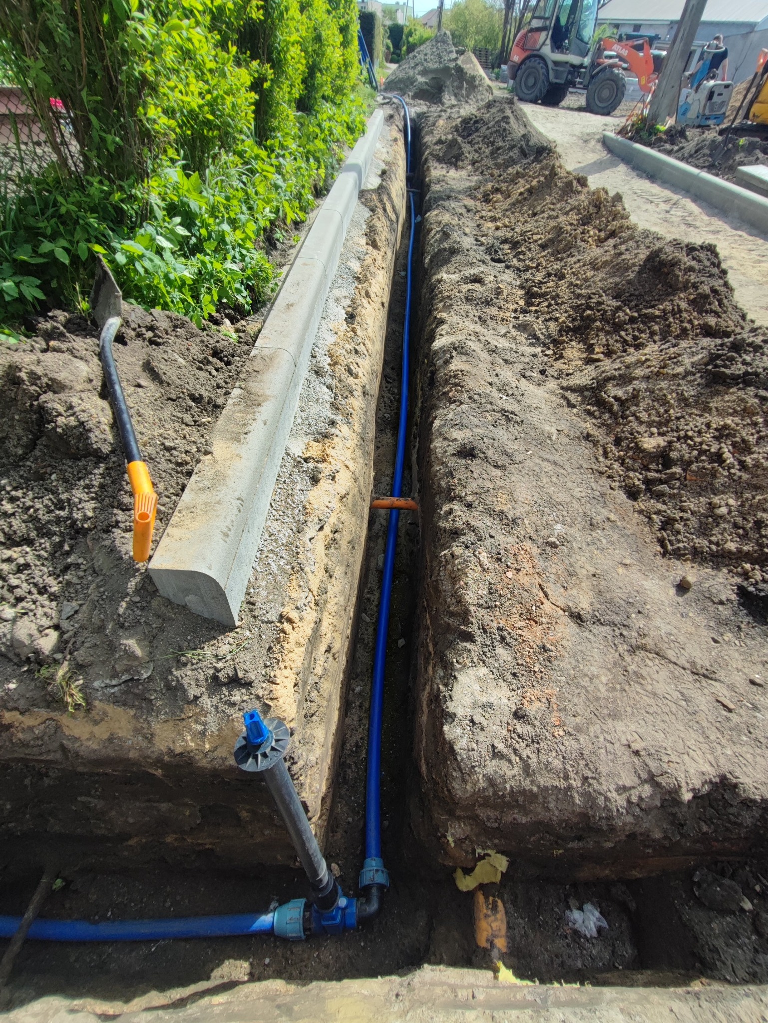 Wkop pod niebieskie rury wodociągowe w ziemi, obok ułożony krawężnik. W tle sprzęt budowlany. Widok z góry na wykop.