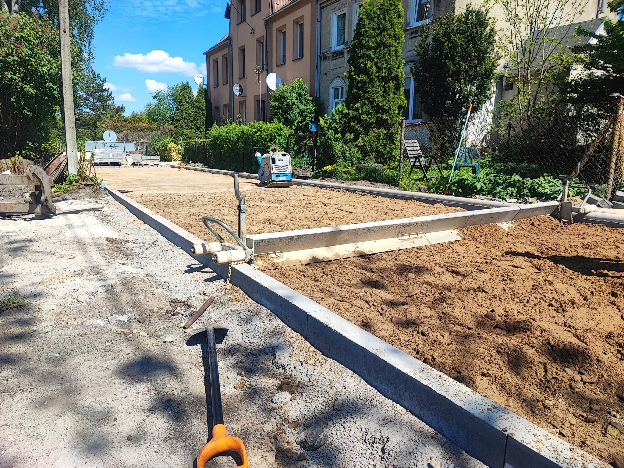 Przygotowanie podłoża pod kostkę brukową w Świdwinie. Obrzeża ułożone, teren wyrównany, zagęszczarka widoczna w tle. Łopata wbita w ziemię na pierwszym planie.