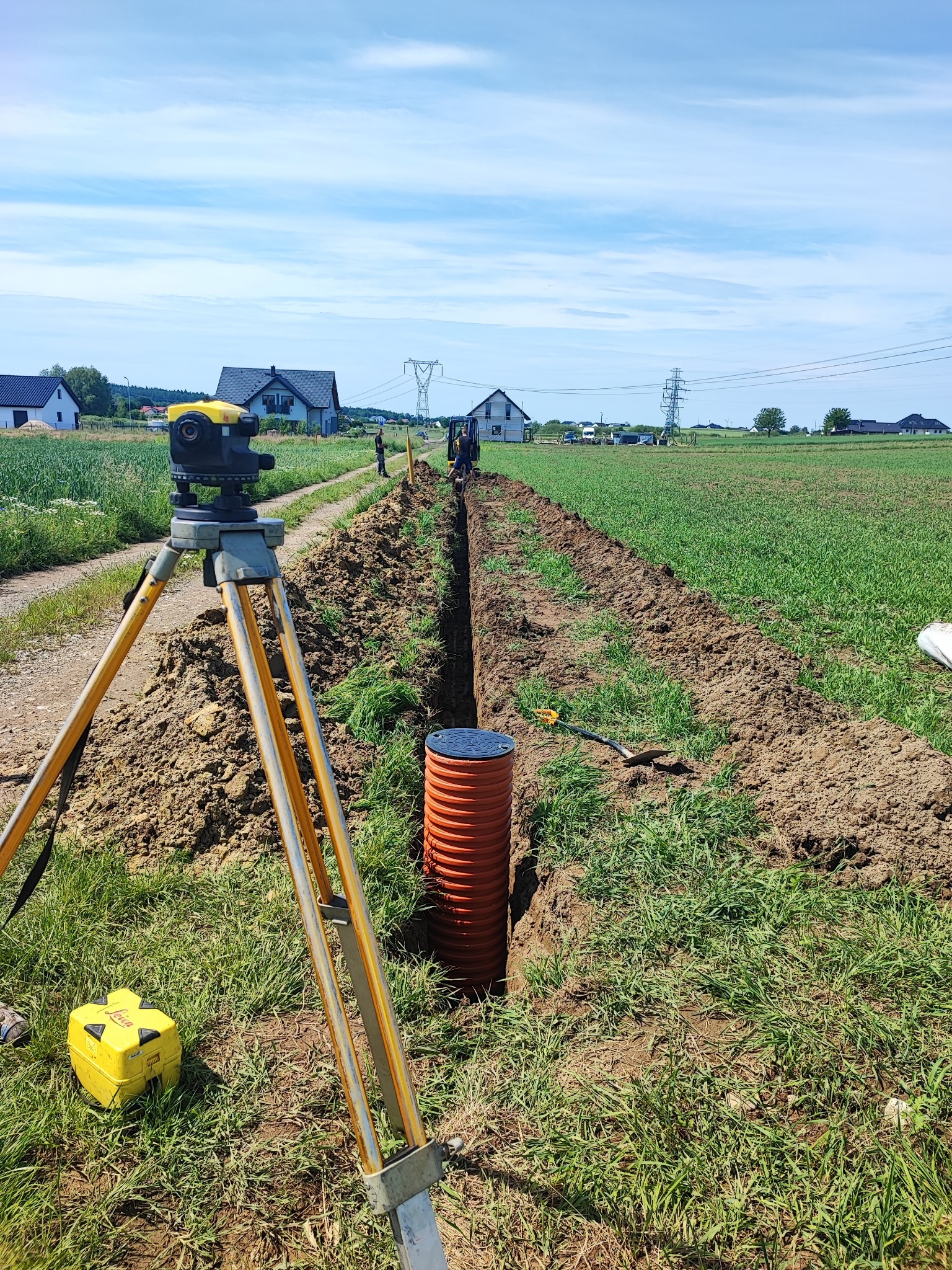 Wykop pod instalacje w terenie wiejskim, widoczny pomarańczowy rurociąg i niwelator Leica na statywie. W tle domy i błękitne niebo.