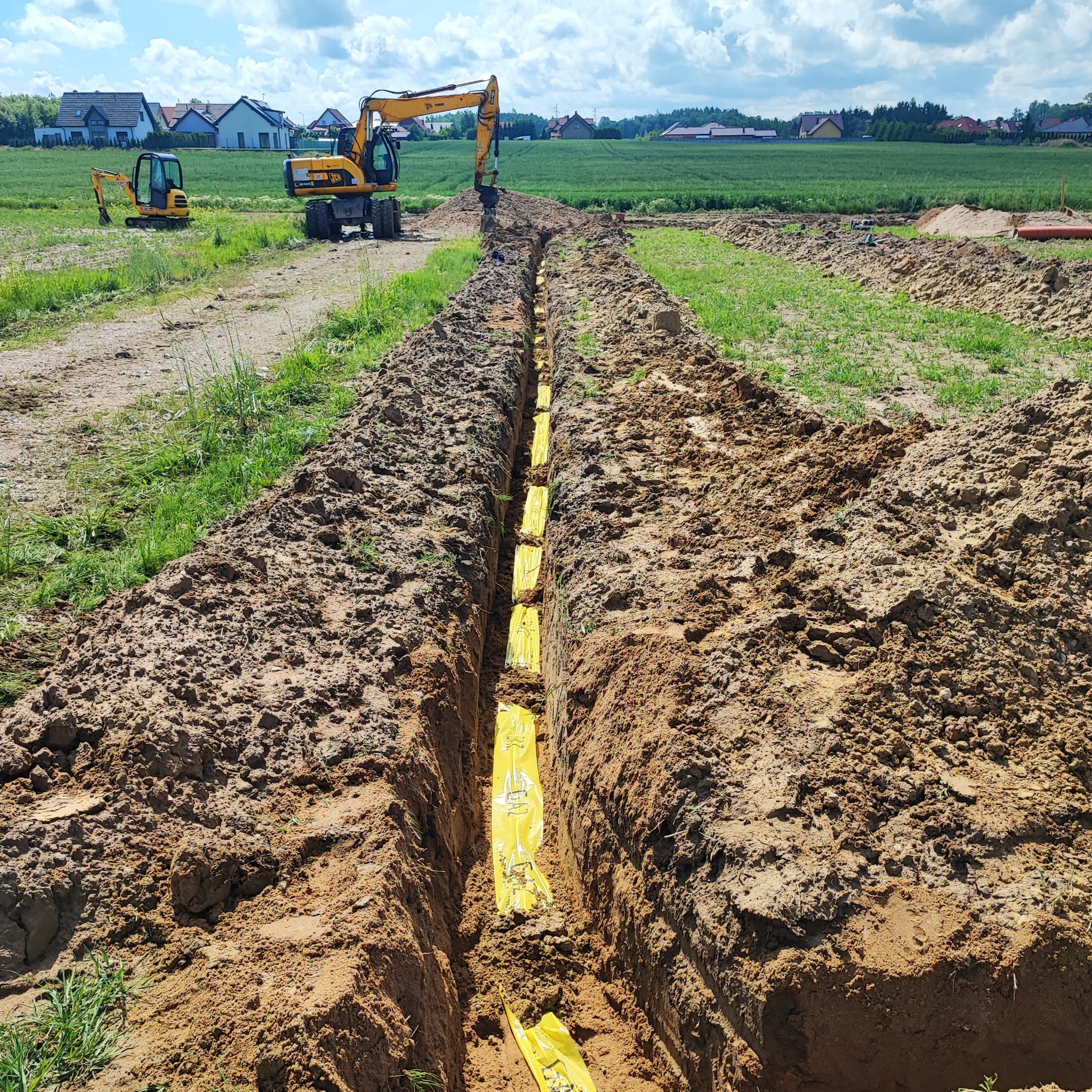 Wykop pod instalacje z żółtą folią zabezpieczającą, widoczne dwie koparki na tle zielonego pola i zabudowań. Wykop świeży, ziemia wilgotna, słoneczny dzień.