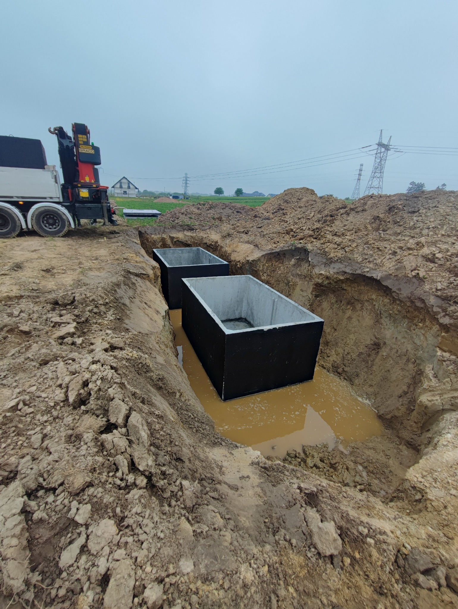 Montaż betonowych zbiorników w wykopie, widoczny dźwig samochodowy. Prace ziemne, woda w wykopie, pochmurne niebo. Dwa zbiorniki w ziemi.