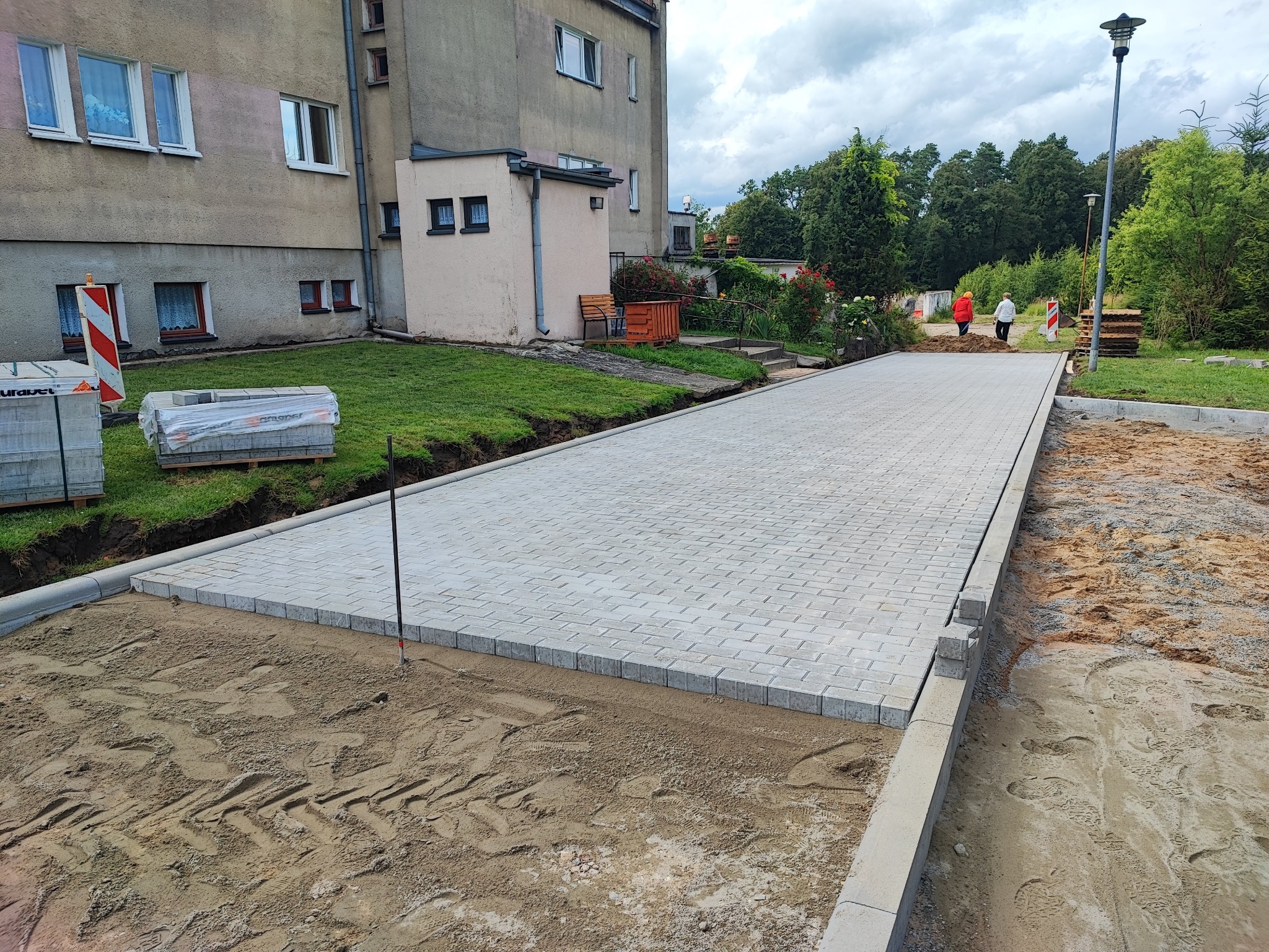Ułożona, szara kostka brukowa tworząca chodnik przed budynkiem mieszkalnym. Częściowo utwardzony teren z widocznymi śladami opon. Paleta kostki brukowej w tle.