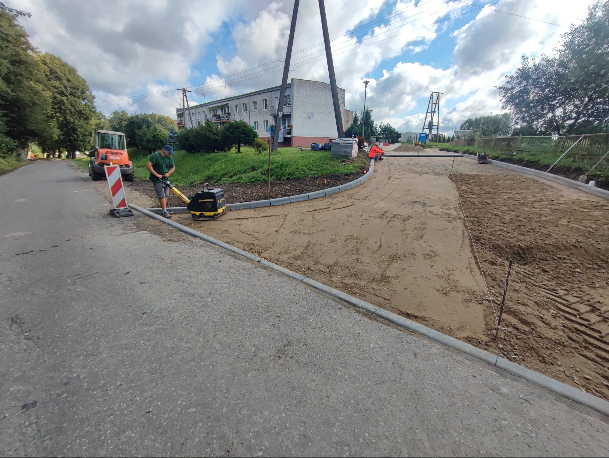 Prace ziemne: zagęszczarka w akcji na przygotowanym podłożu, obok ułożone krawężniki. W tle budynek mieszkalny i maszyna budowlana. Etap przygotowania podłoża pod kostkę.