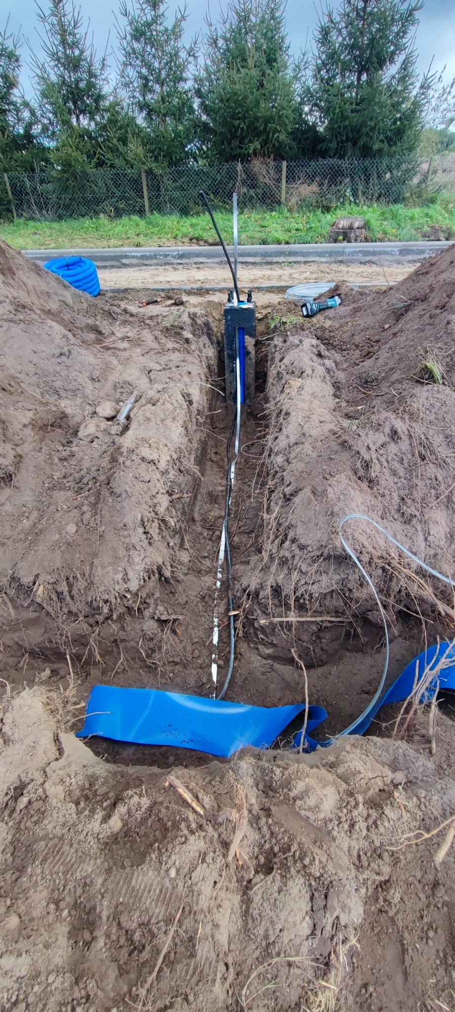 Wąski wykop w ziemi z widoczną instalacją w czarnej obudowie, niebieskie rury w tle i na pierwszym planie. Prace ziemne w trakcie realizacji.