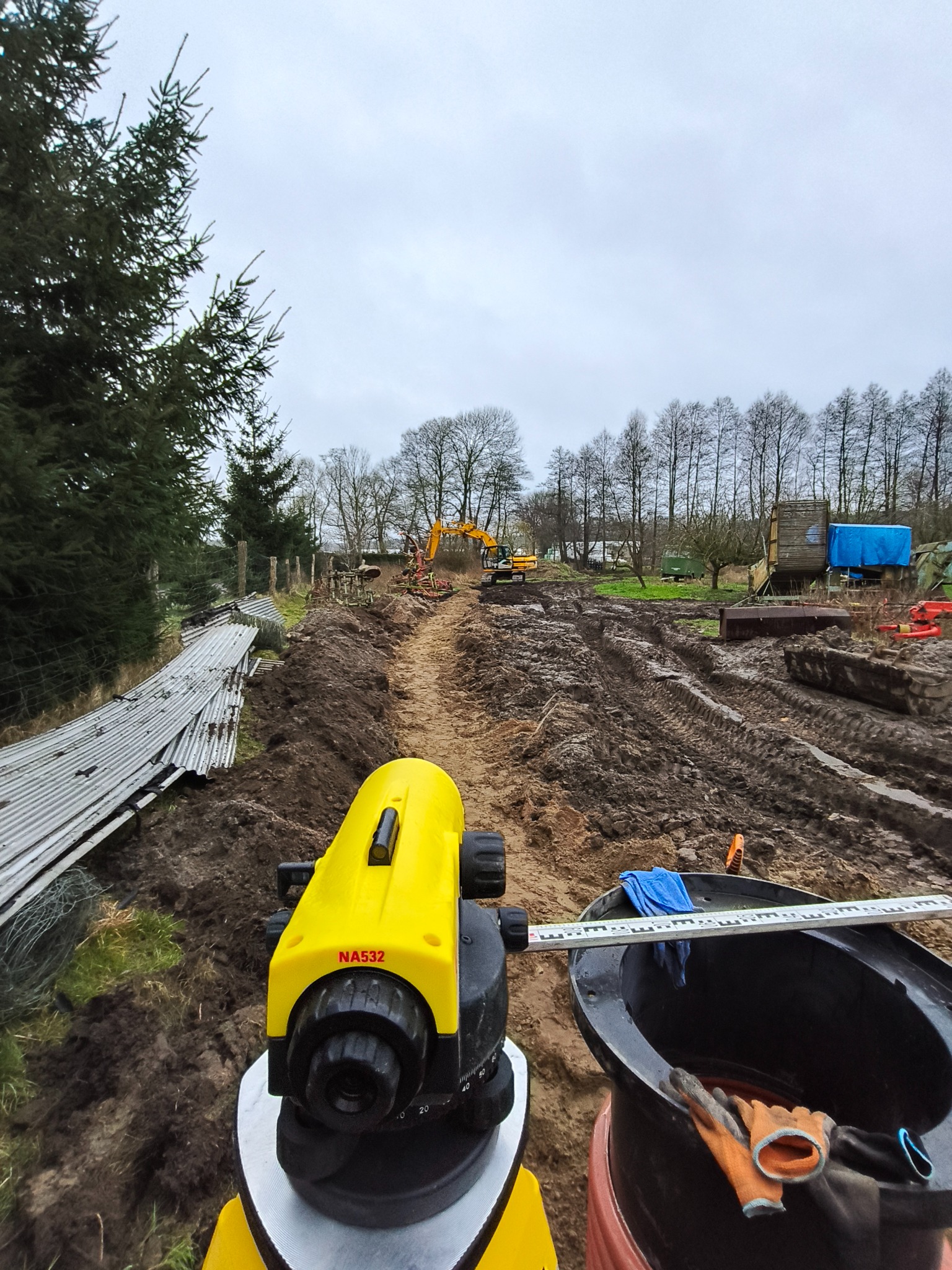 Żółty niwelator NA532 na pierwszym planie, w tle wykop pod instalacje z pracującą koparką. Widoczne narzędzia i elementy infrastruktury na placu budowy.