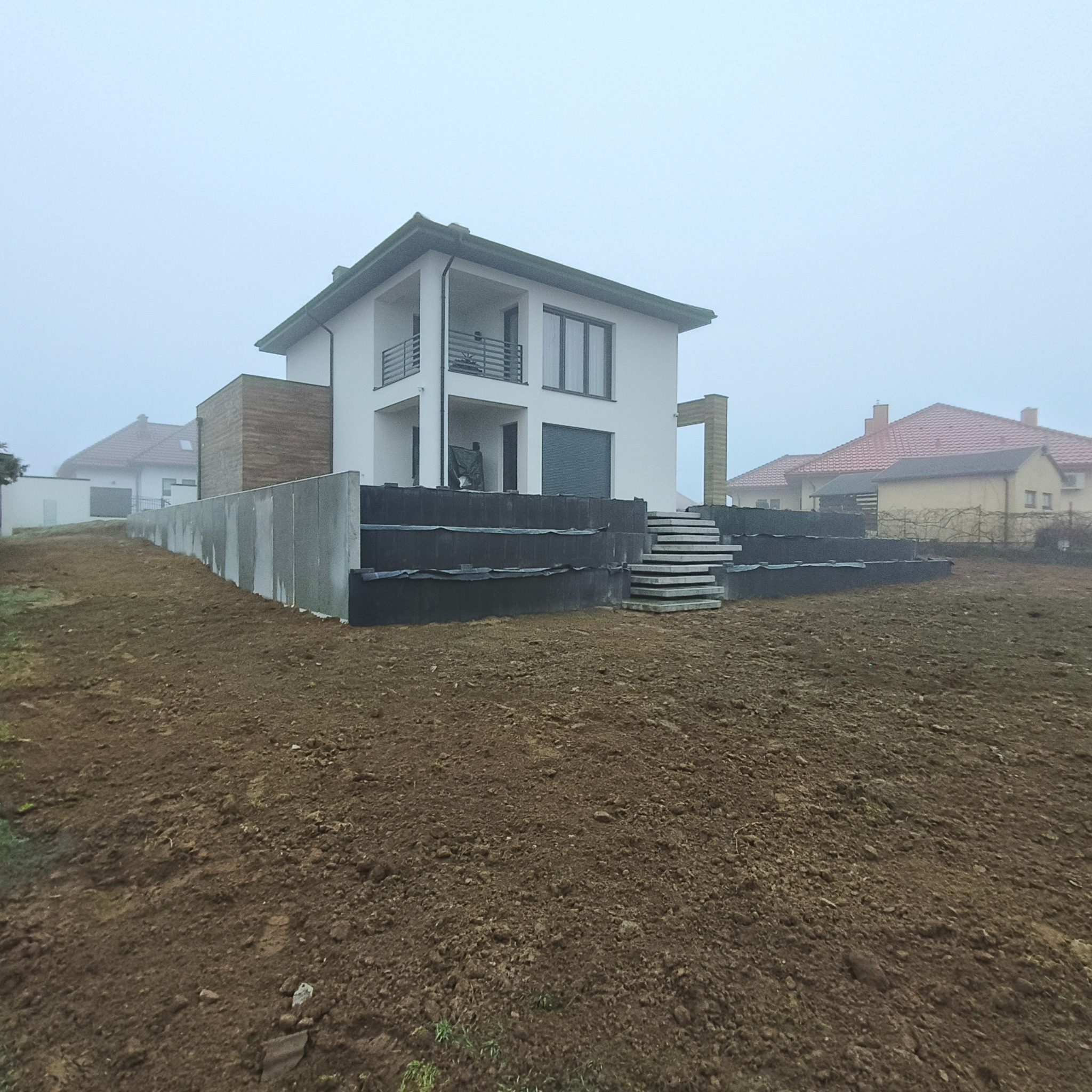 Nowoczesny dom z betonowymi schodami i murem oporowym na nierównym terenie, przygotowany pod dalsze prace ziemne w pochmurny dzień.