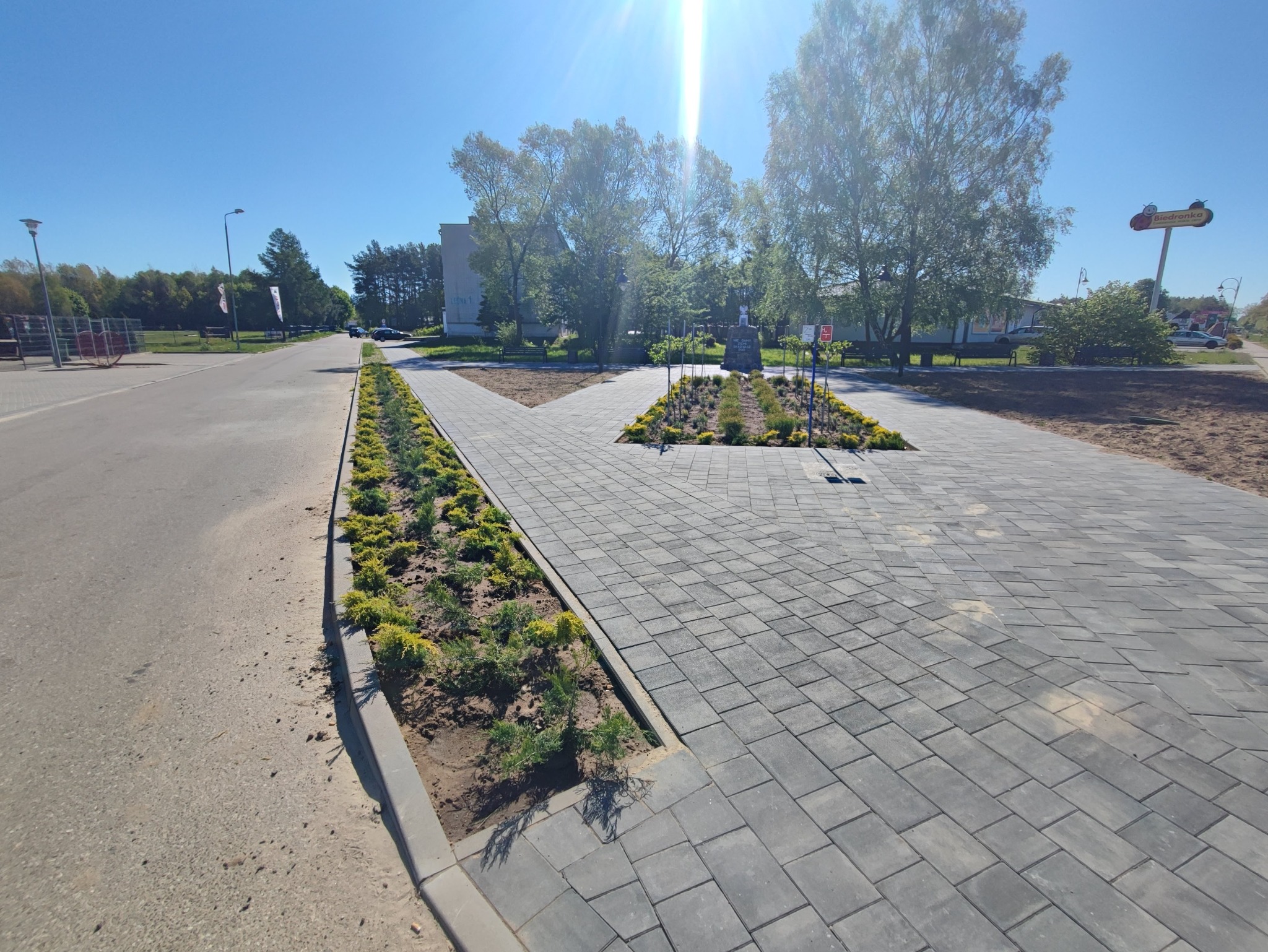 Szara kostka brukowa ułożona na placu z rabatami zieleni. Słoneczny dzień, widok na pomnik i drzewa w tle. Geometryczny wzór kostki, świeżo posadzone rośliny.