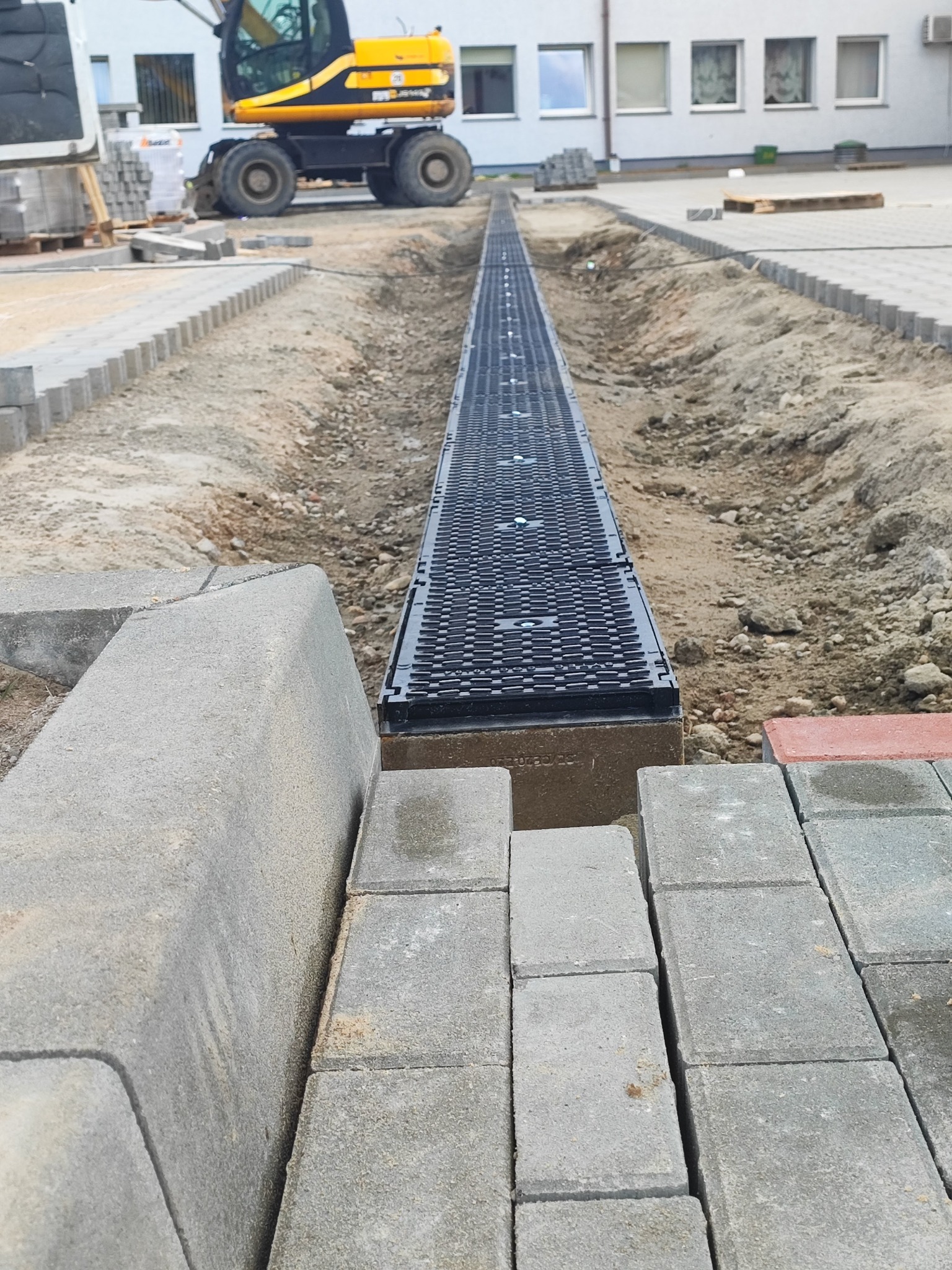 Montaż odwodnienia liniowego z kratką na podsypce betonowej, przygotowanie do układania kostki brukowej. W tle koparka kołowa i budynek.