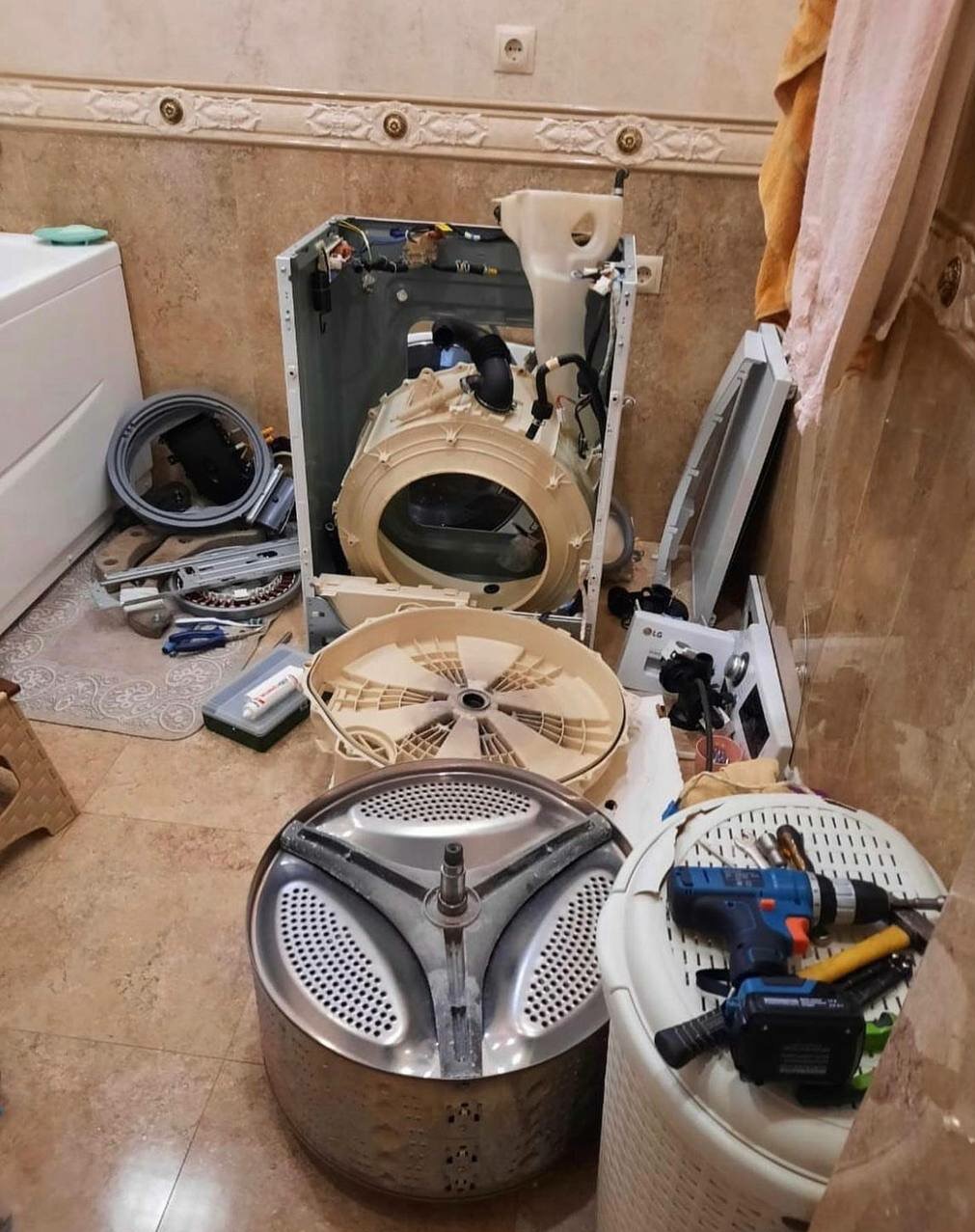 Rozłożona pralka LG w trakcie naprawy. Widoczne elementy bębna, obudowy i narzędzia, w tym wkrętarka. Beżowe płytki w tle.