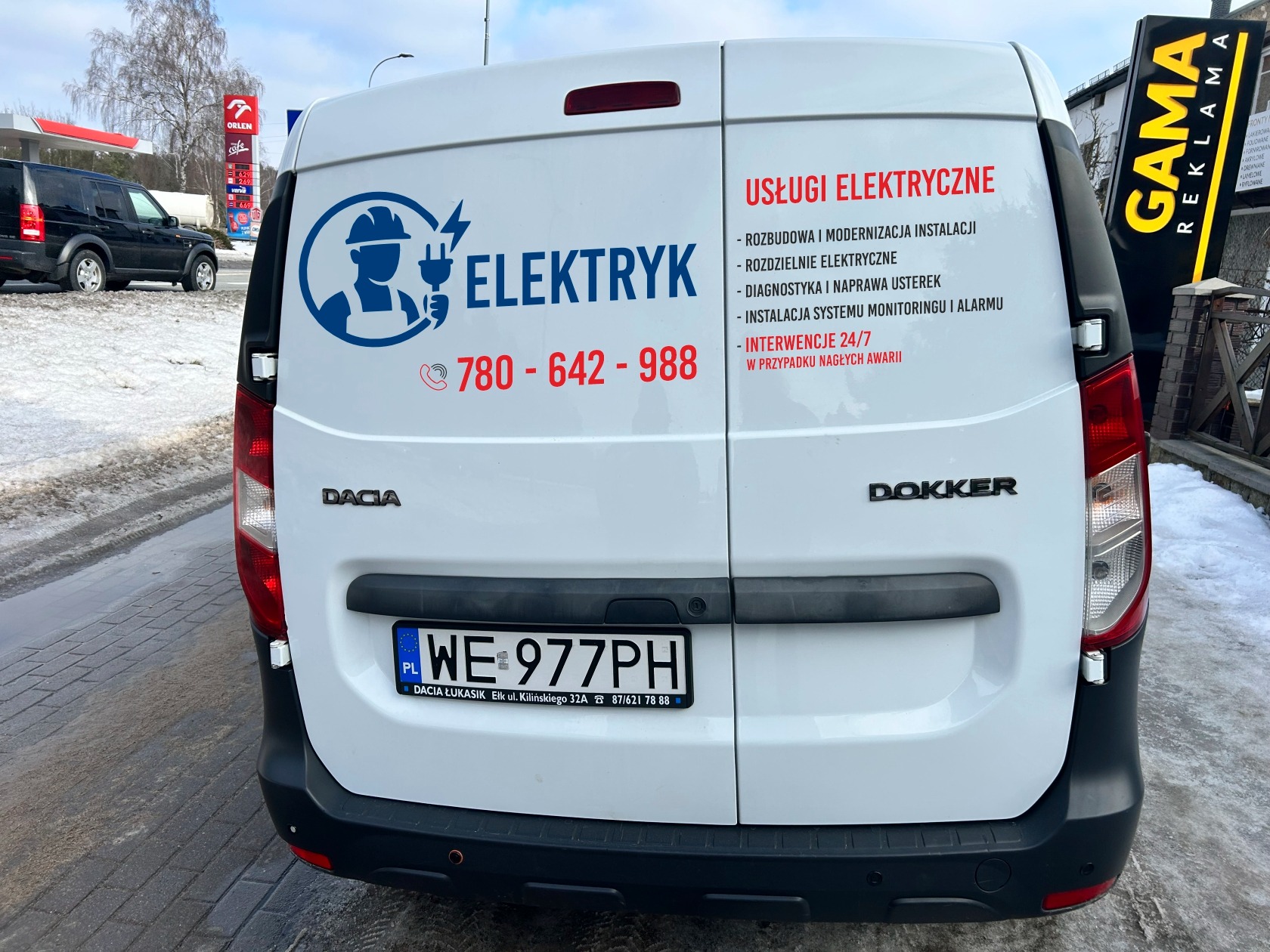Biały samochód dostawczy Dacia Dokker z reklamą usług elektrycznych, numer telefonu i logo firmy. Widok z tyłu na tle stacji benzynowej Orlen i reklamy Gama.